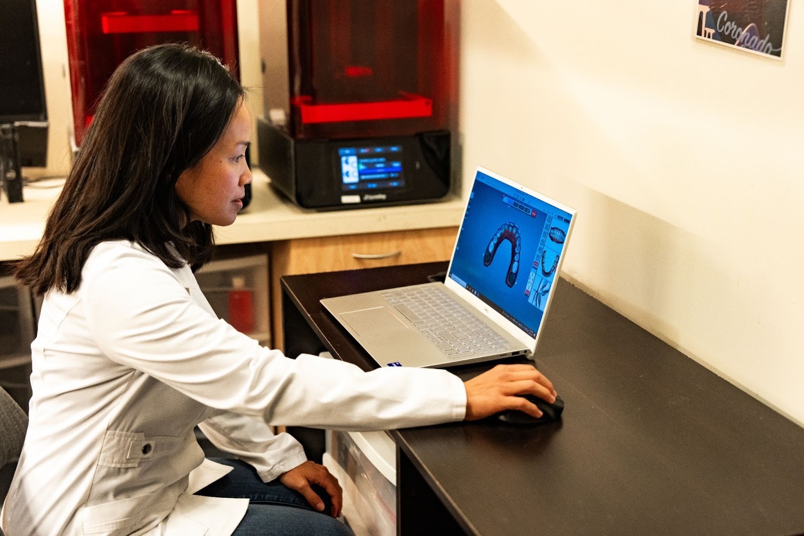 Orthodontist in a lab coat using a laptop to design a retainer, with 3D printer in background, emphasizing retainer fitting and maintenance.